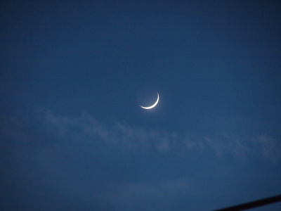 [The early night sky is slate blue. A lower right sliver of moon is visible and very white while the rest of the moon circle in a different shade of slate blue and thus apparent.]